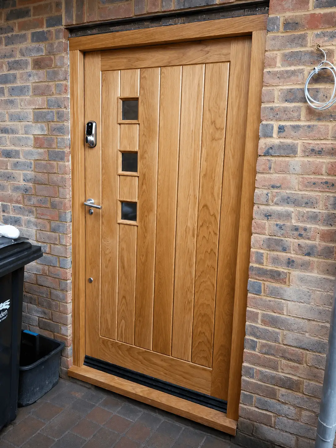 Contemporary oak front door with vertical boarding and three square glazed lights, a modern lever handle and keypad, in a brick surround