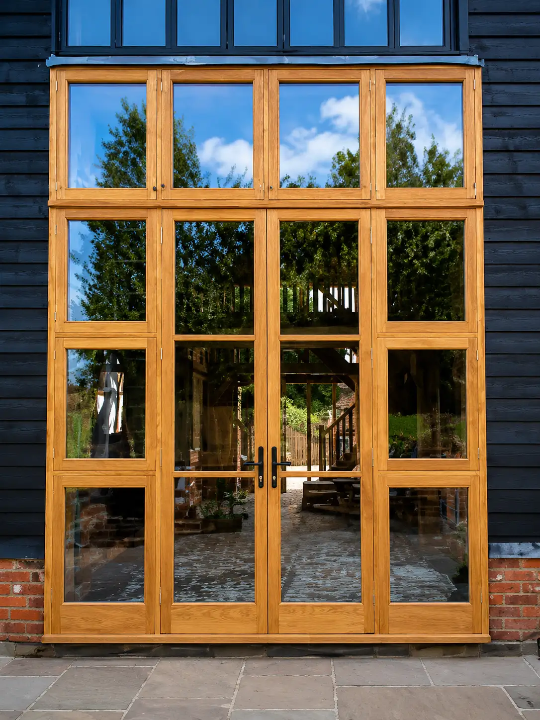 Oak fully-glazed French double doors with a multi-pane glazed transom screen above, set into black weatherboarding over a red-brick base