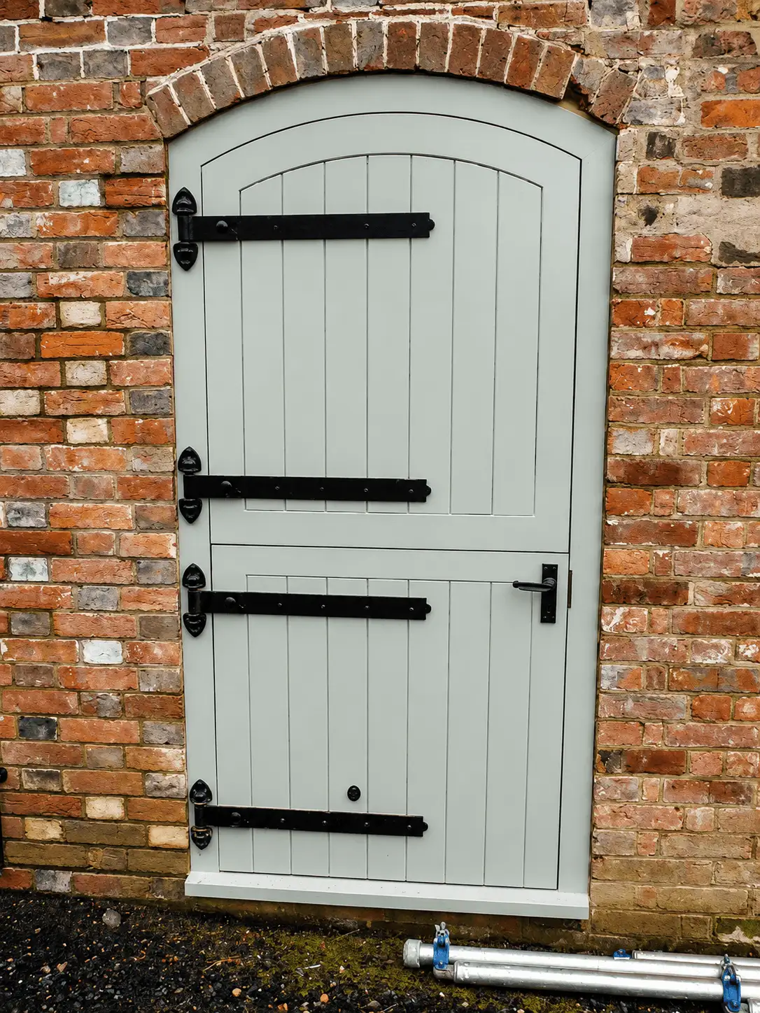 Sage-green painted arched stable door with black strap hinges, set in a weathered brick opening
