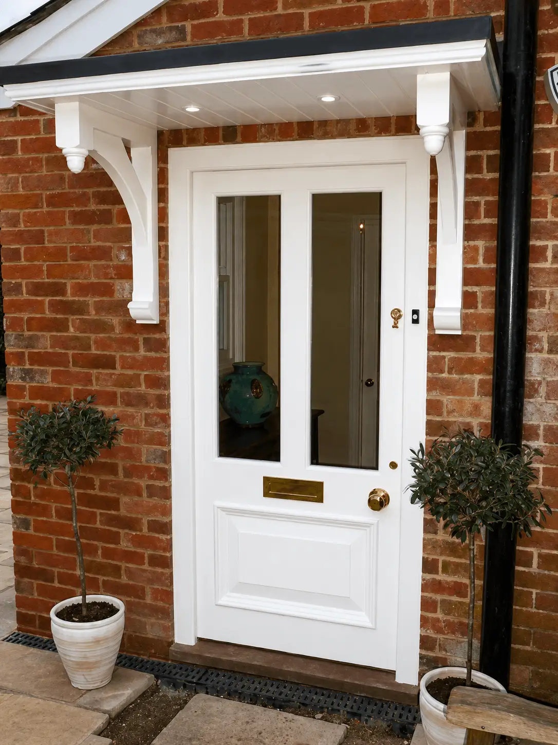 White painted half-glazed front door with brass furniture, under a timber porch canopy in a brick surround