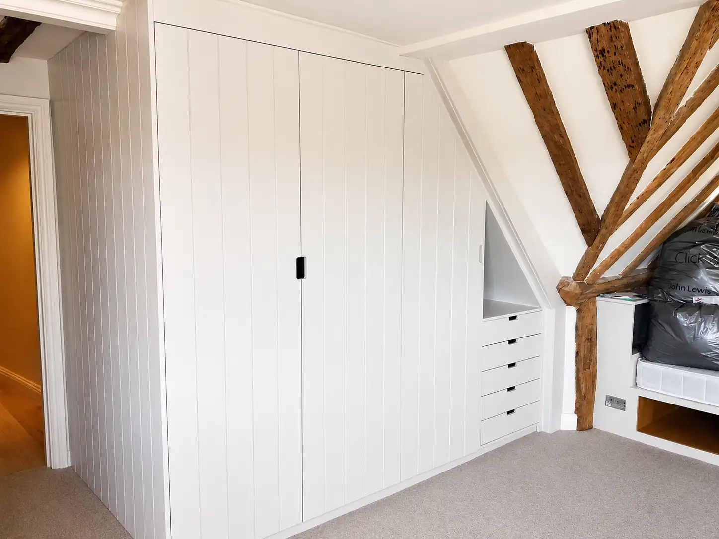 White boarded fitted wardrobes in a beamed loft bedroom, angled around the eaves with a drawer bank under the slope beside an exposed oak truss
