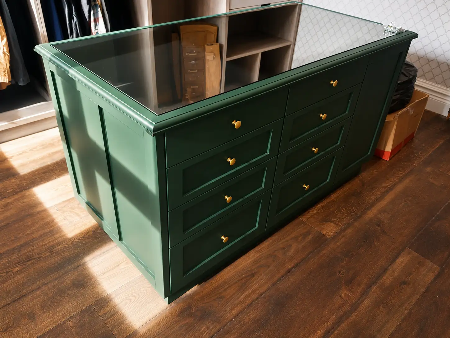 Bottle-green panelled dressing-room island with a glass top, brass knobs and banks of drawers in a walk-in wardrobe